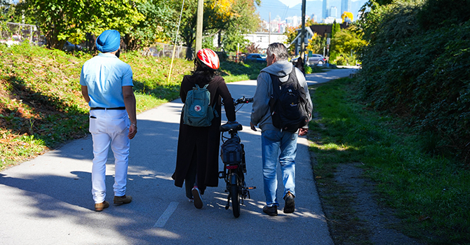 Three people walking along the Arbutus Greenway, one person has a bike. Three people walking along the Arbutus Greenway, one person has a bike.