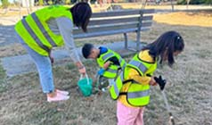 One adult and two children in high-visibility vests using litter pickers and buckets to clean up grass in a park.  One adult and two children in high-visibility vests using litter pickers and buckets to clean up grass in a park.
