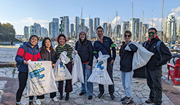 A group of 7 smiling volunteers with neighbourhood cleanup labelled garbage bags standing near the False Creek seawall.  A group of 7 smiling volunteers with neighbourhood cleanup labelled garbage bags standing near the False Creek seawall.