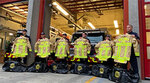 A group of firefighters standing in front of a firetruck, each holding new yellow turnout jackets labeled 'Vancouver Fire.' Black gear bags are placed on the ground in front of them. A group of firefighters standing in front of a firetruck, each holding new yellow turnout jackets labeled 'Vancouver Fire.' Black gear bags are placed on the ground in front of them.